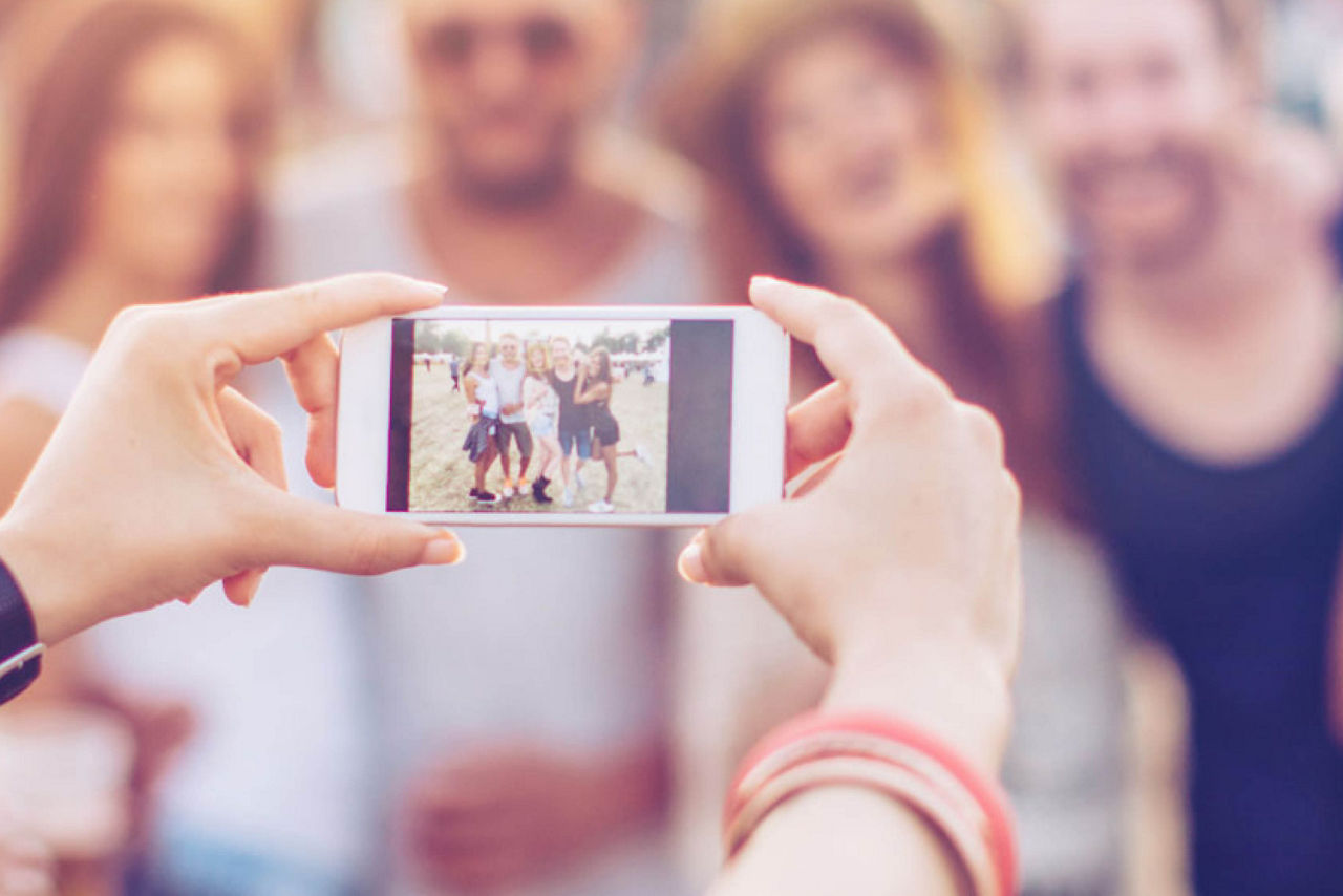 Im Vordergrund halten zwei Hände ein Smartphone, das ein Bild von einer Gruppe junger Menschen macht. Die Gruppe  ist im Hintergrund unscharf zu sehen.