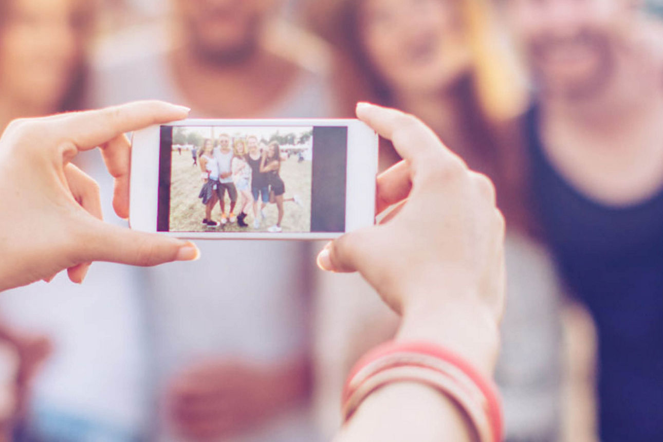 Im Vordergrund halten zwei Hände ein Smartphone, das ein Bild von einer Gruppe junger Menschen macht. Die Gruppe  ist im Hintergrund unscharf zu sehen.