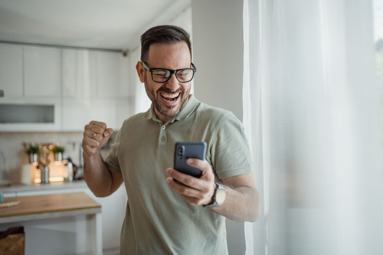 Ein Mann steht in seiner Küche und blickt jubelnd auf sein Smartphone