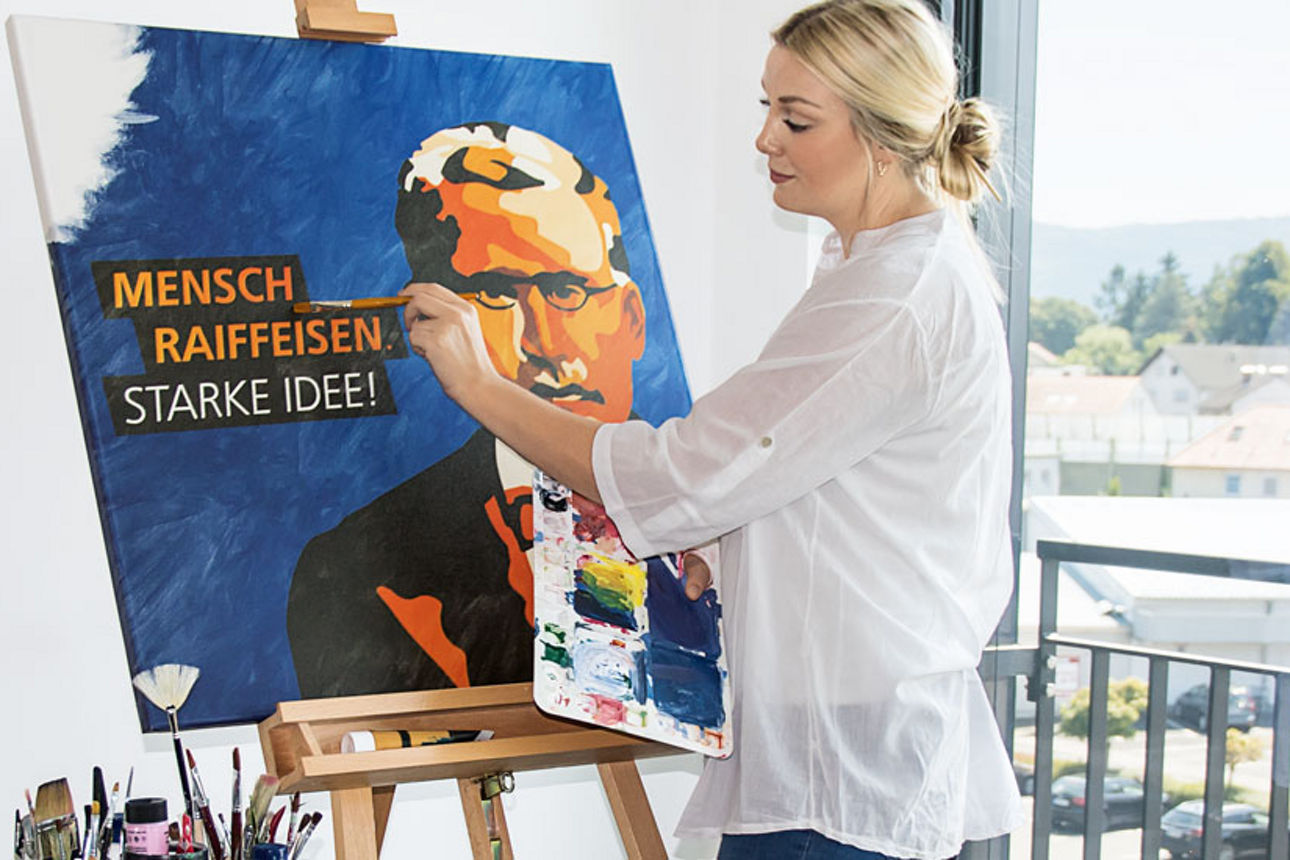 Eine Frau malt an einer Staffelei ein Porträt mit dem Text ‚Mensch Raiffeisen – Starke Idee‘. Sie steht in einem hellen Raum 