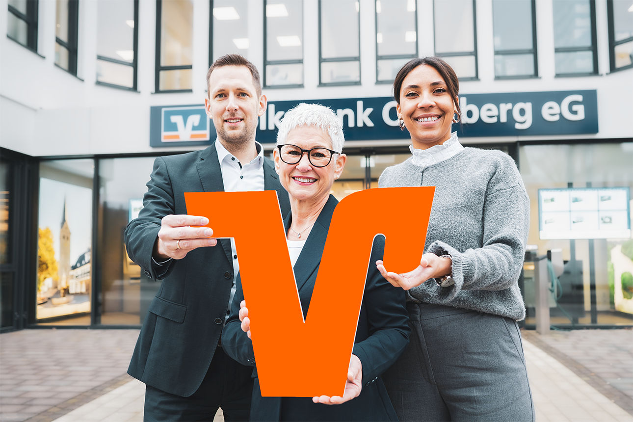 Drei Mitarbeitende stehen lächelnd vor einer Volksbank-Filiale und halten gemeinsam ein großes orangefarbenes ‚V‘ als Symbol der Bank.