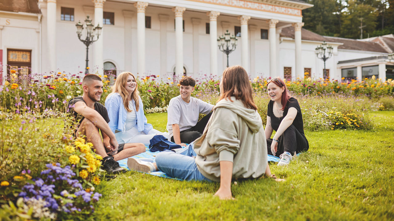 Fünf junge Menschen sitzen auf einer Wiese in einem Park und unterhalten sich lächelnd.