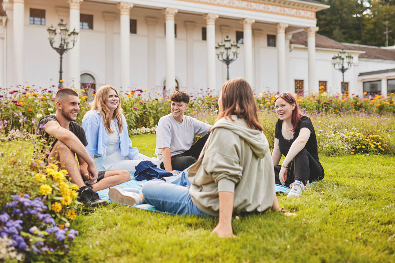 Fünf junge Menschen sitzen auf einer Wiese in einem Park und unterhalten sich lächelnd.