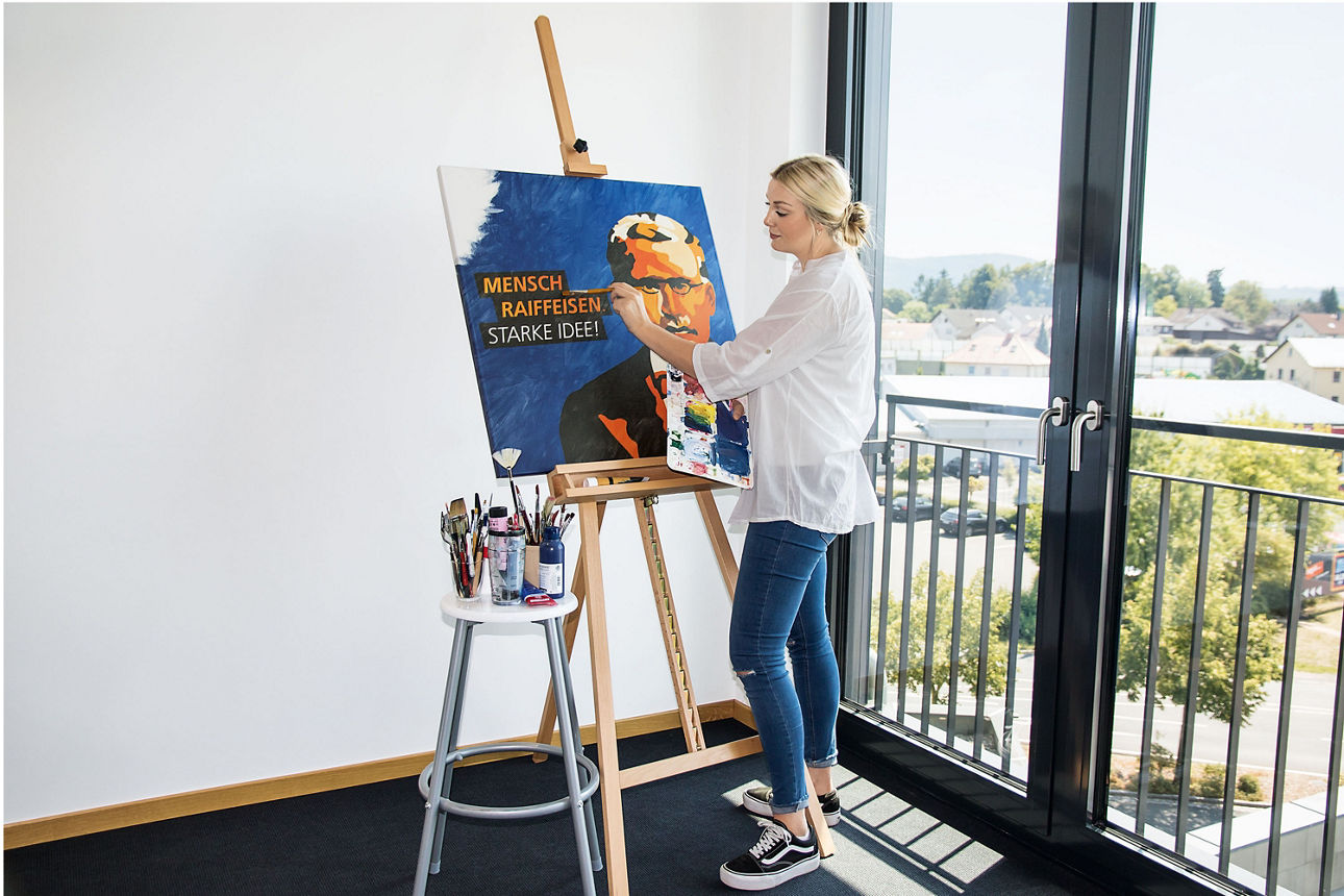 Eine Frau malt an einer Staffelei ein Porträt mit dem Text ‚Mensch Raiffeisen – Starke Idee‘. Sie steht in einem hellen Raum mit Blick nach draußen.
