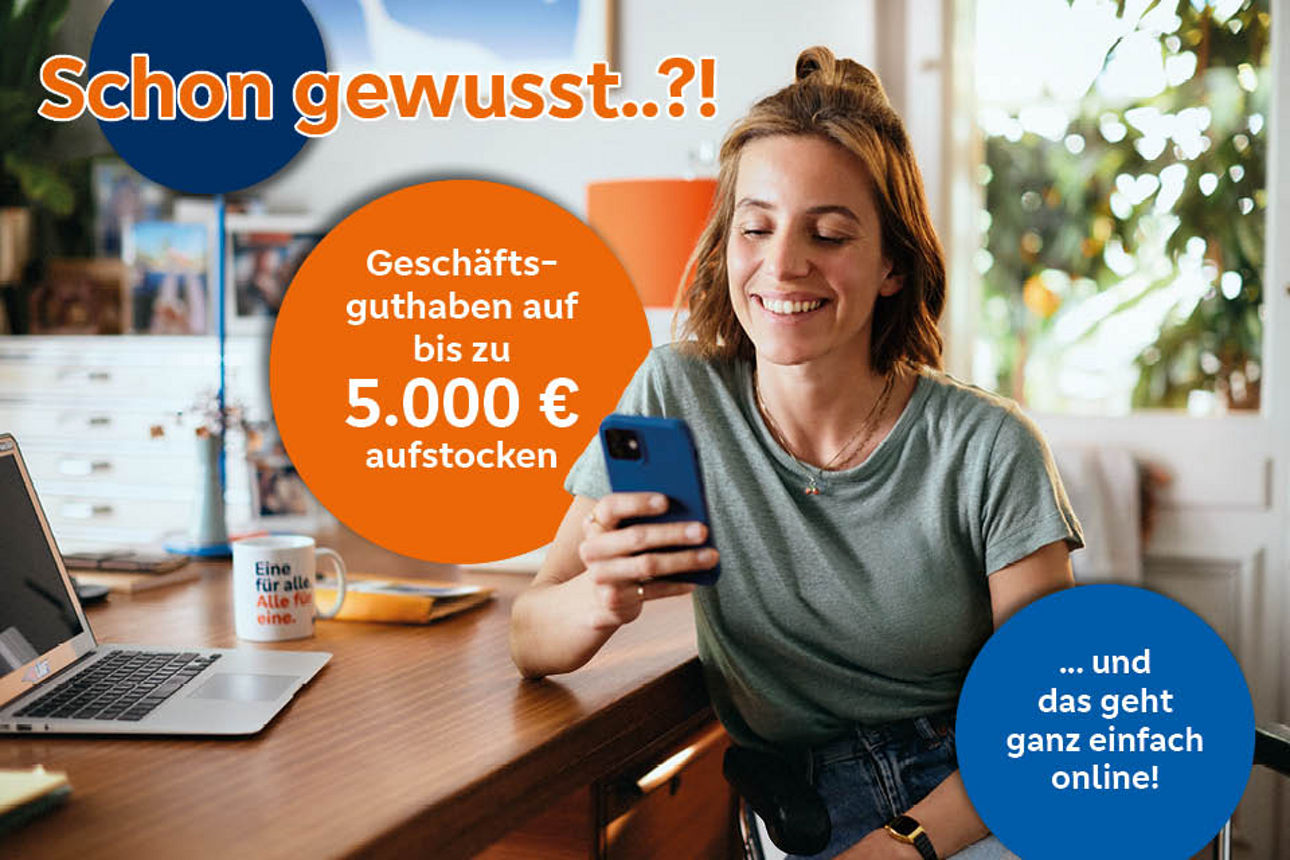 Junge Frau sitzt am Tisch mit Laptop und schaut auf ihr Smartphone