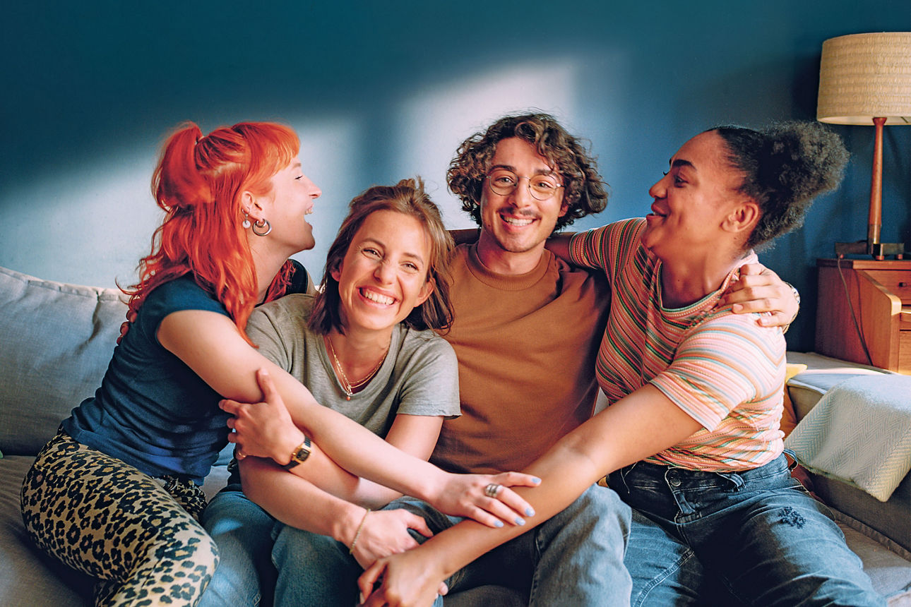 Vier junge Menschen sitzen auf einer Couch, nehmen sich in den Arm und lächeln.