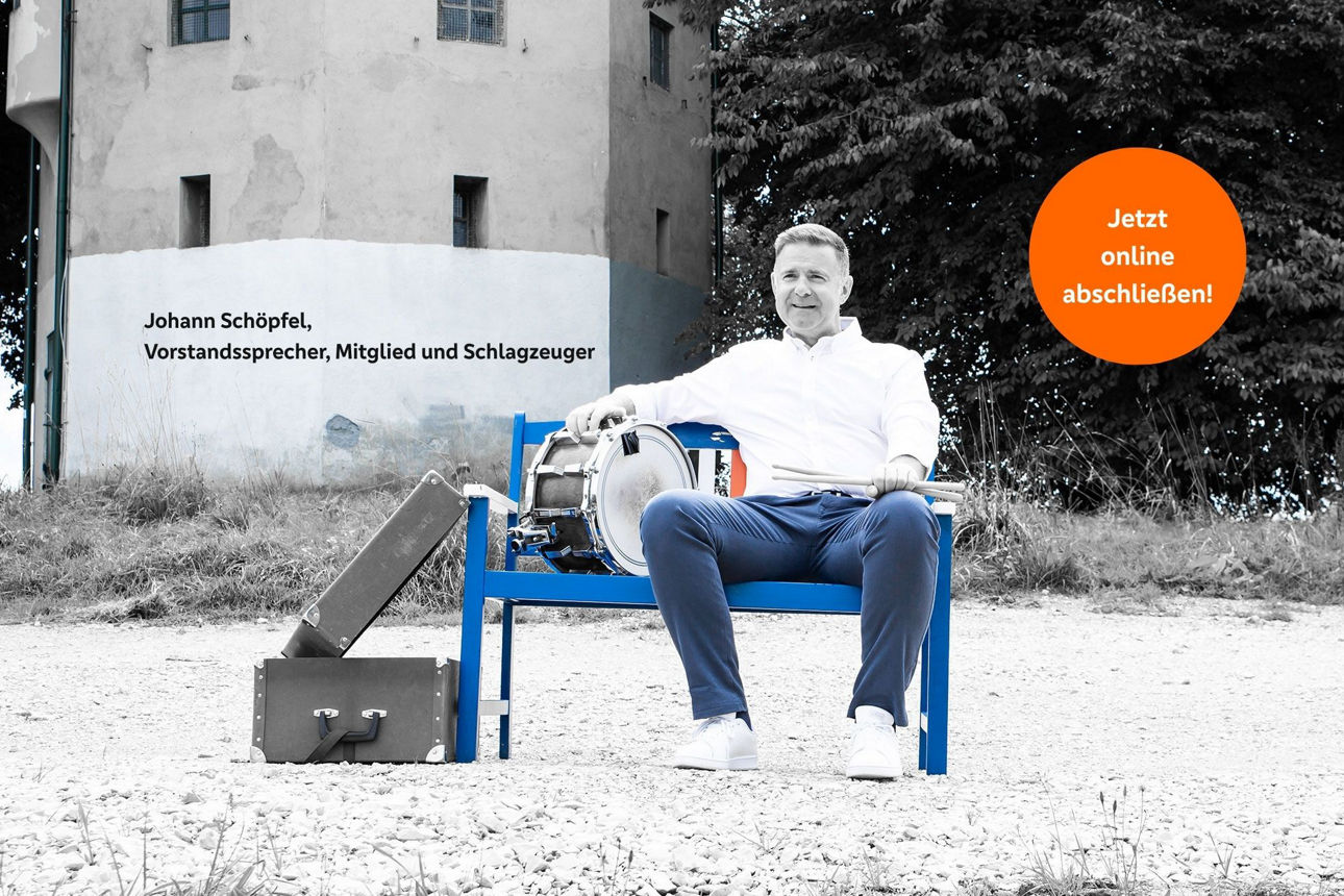Mann sitzt auf einer Holzbank mit einer Trommel vor einem Gebäude im Grünen