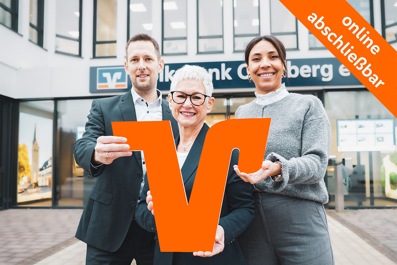 Drei Mitarbeitende stehen lächelnd vor einer Volksbank-Filiale und halten gemeinsam ein großes orangefarbenes ‚V‘ als Symbol der Bank.