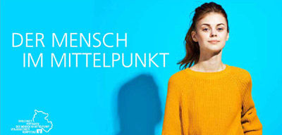 Eine Frau trägt einen gelben Pullover, auf dem der Satz "der mensch im mittelepunkt" prangt. 