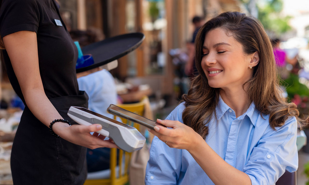 Junge Frau sitzt im Außenbereich eines Restaurants und hält ihr Smartphone zur Bezahlung an ein Kartenlesegerät.