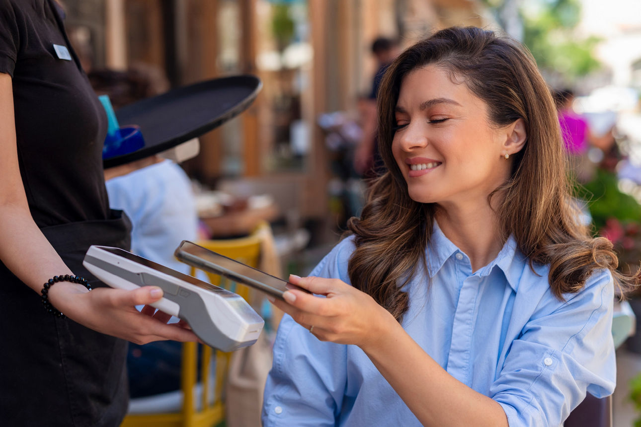 Junge Frau sitzt im Außenbereich eines Restaurants und hält ihr Smartphone zur Bezahlung an ein Kartenlesegerät.