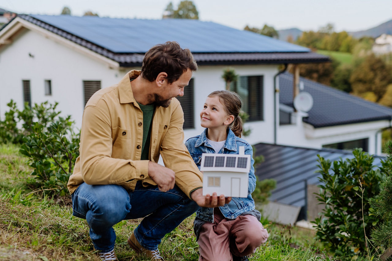 Ein Vater und seine kleine Tochter hocken draußen mit einem Modellhaus in der Hand und lächeln sich an.