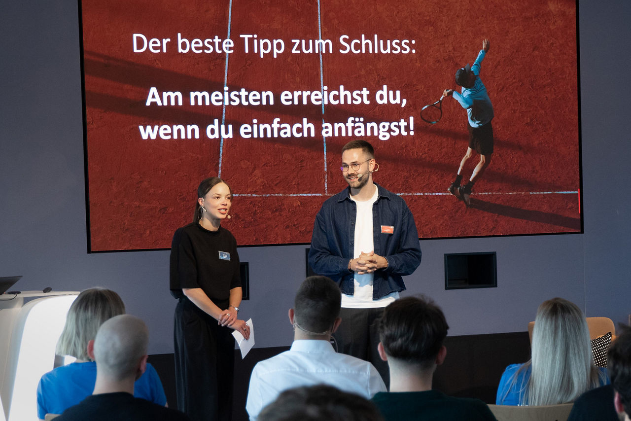 Die beiden Vortragenden stehen vor Publikum vor einer Leinwand mit dem Schlussimpuls: „Am meisten erreichst du, wenn du einfach anfängst!“