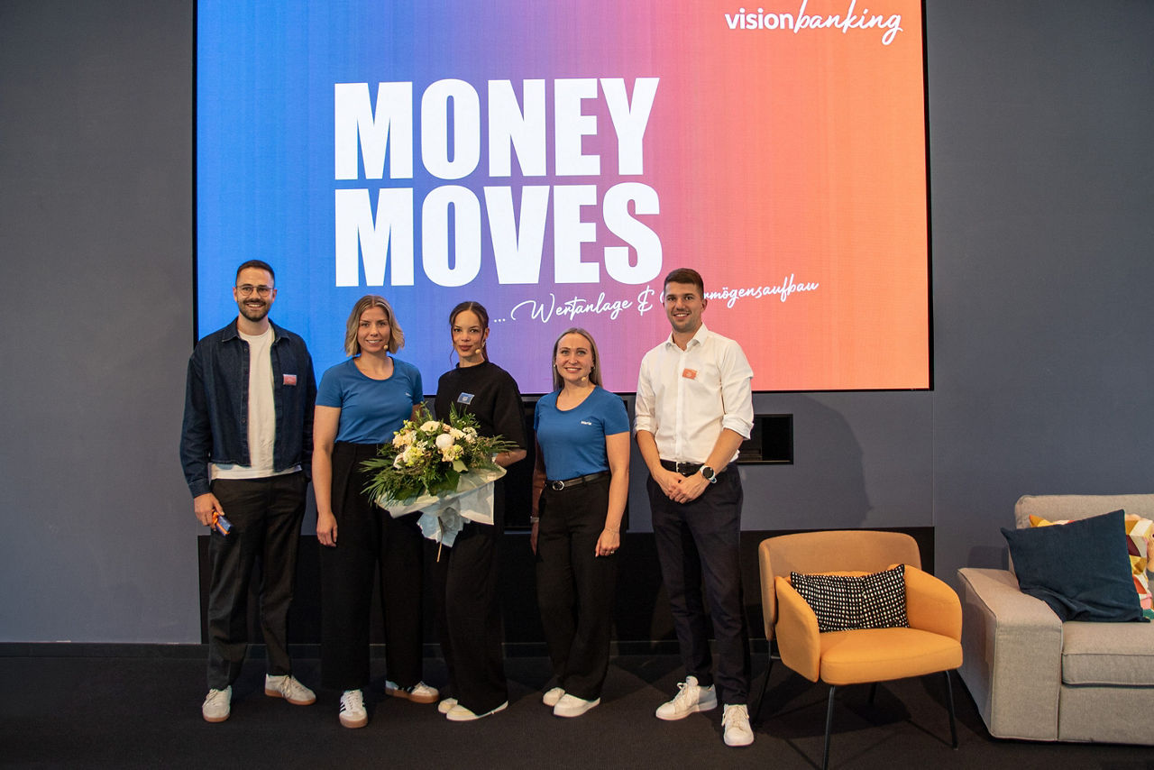 Fünf Personen stehen vor einer Leinwand mit der Aufschrift „Money Moves“; eine Person hält einen Blumenstrauß.