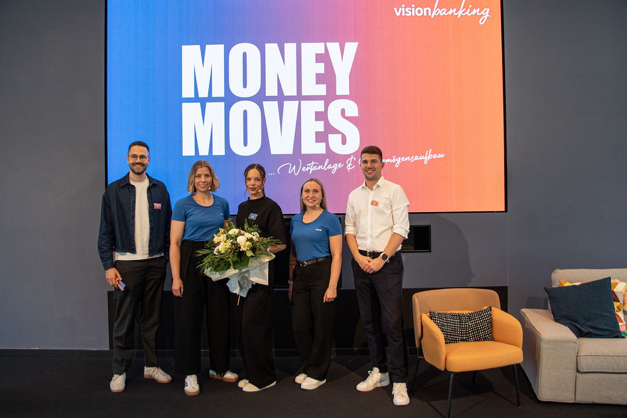 Fünf Personen stehen vor einer Leinwand mit der Aufschrift „Money Moves“; eine Person hält einen Blumenstrauß.