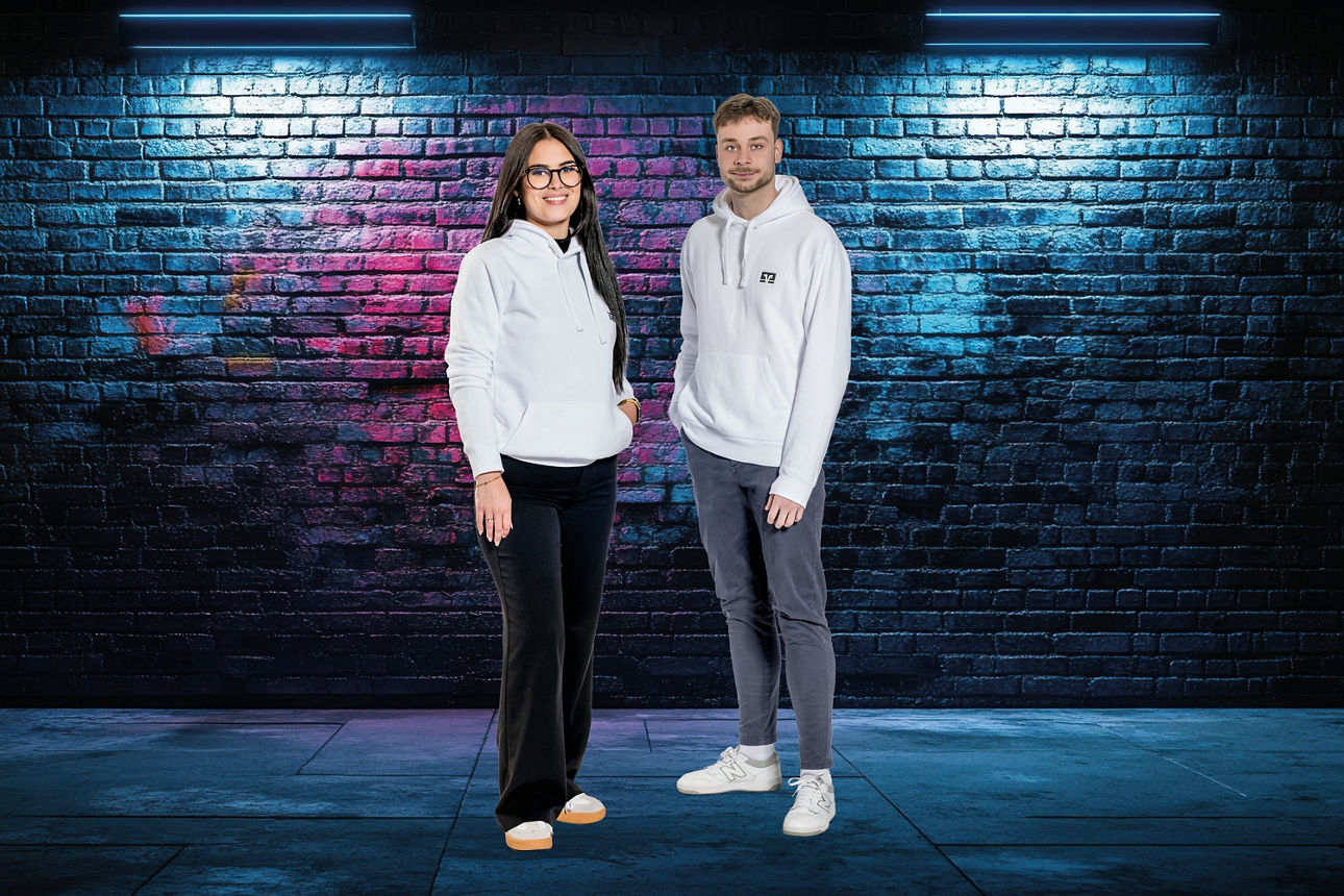 Person in weißem Kapuzenpullover, dunkler Hose und weißen Sneakers vor einer mit buntem Graffiti besprühten Backsteinwand unter zwei Wandlampen stehend