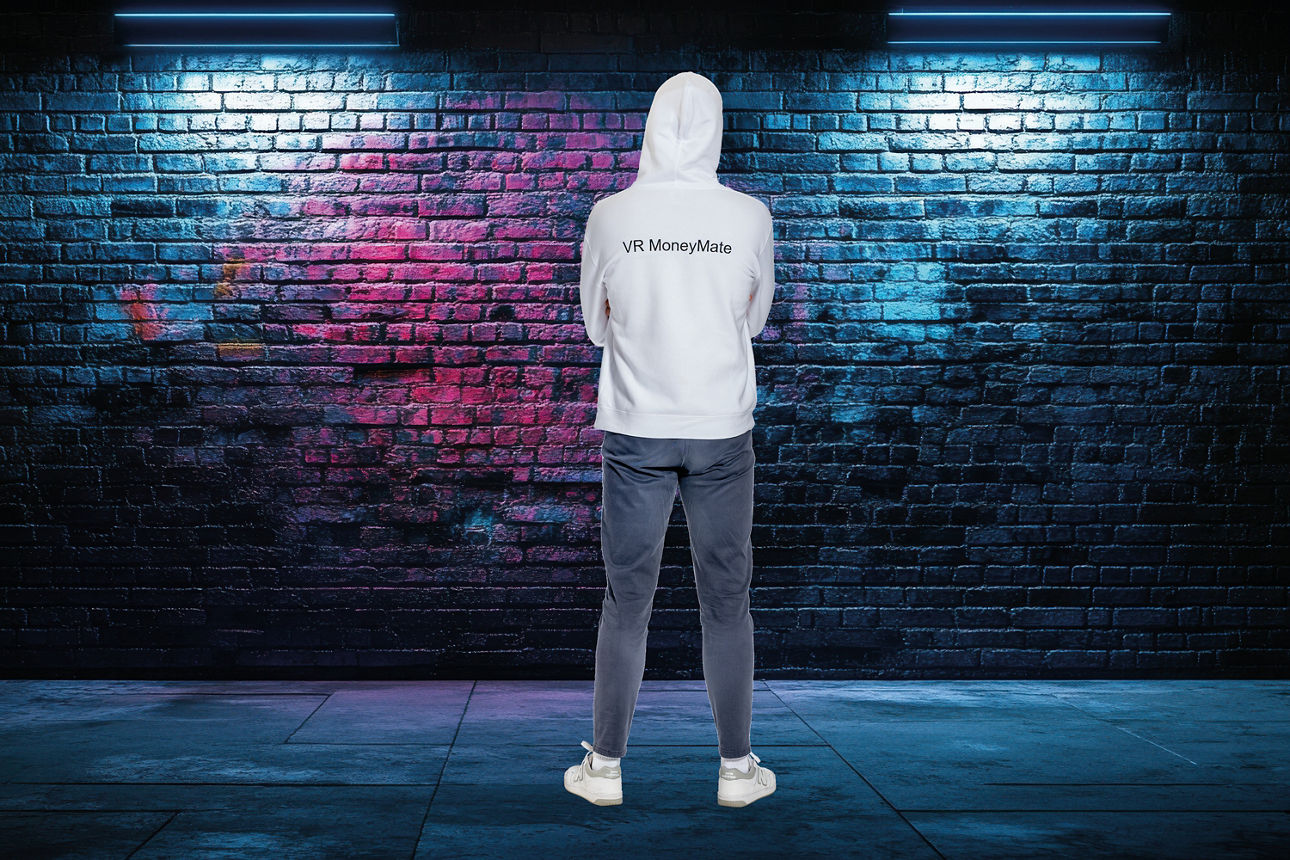 Person in weißem Kapuzenpullover, dunkler Hose und weißen Sneakers vor einer mit buntem Graffiti besprühten Backsteinwand unt