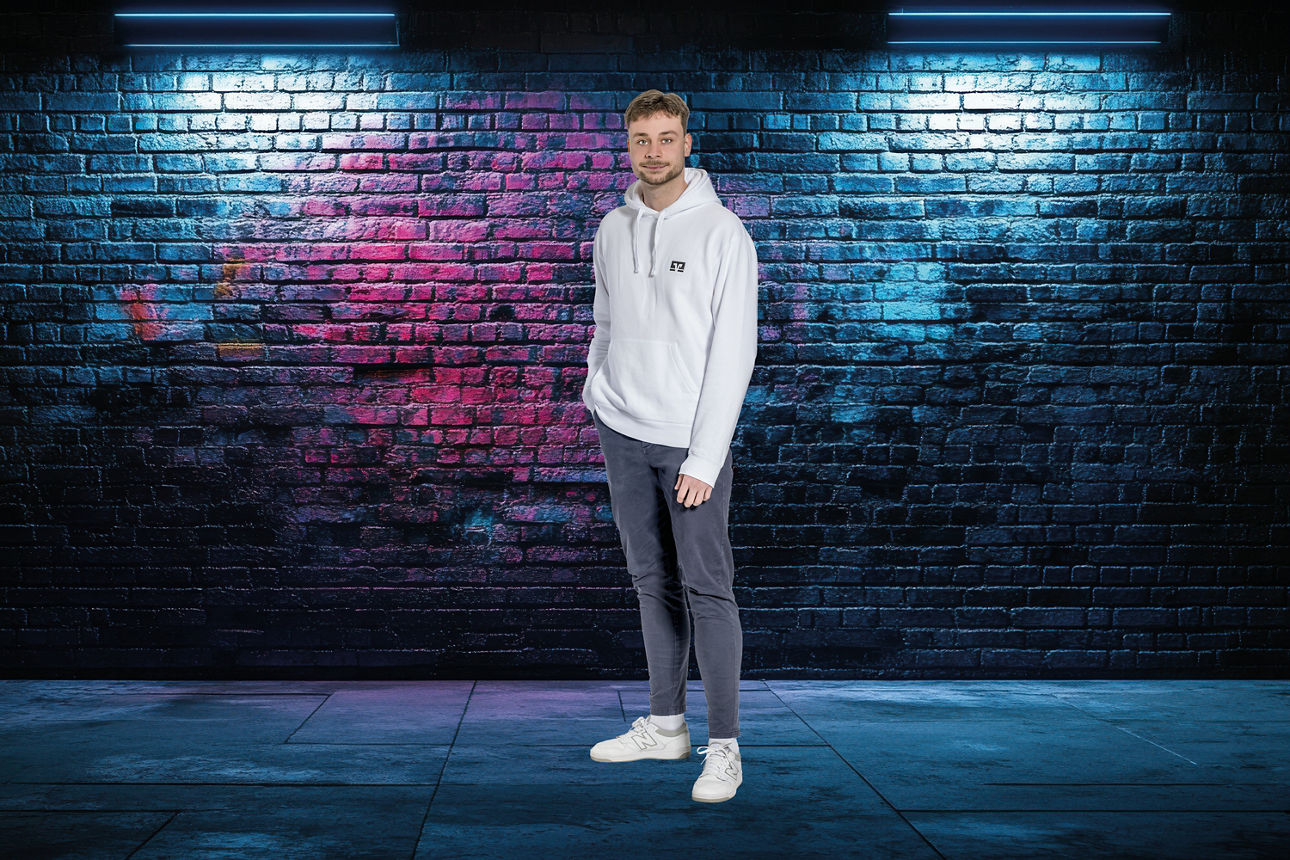 Person in weißem Kapuzenpullover, dunkler Hose und weißen Sneakers vor einer mit buntem Graffiti besprühten Backsteinwand unter zwei Wandlampen stehend