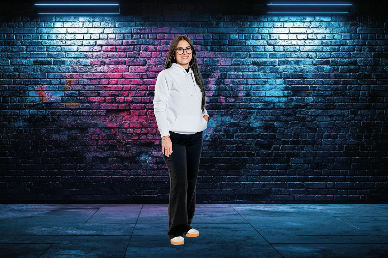 Person in weißem Kapuzenpullover, dunkler Hose und weißen Sneakers vor einer mit buntem Graffiti besprühten Backsteinwand unt