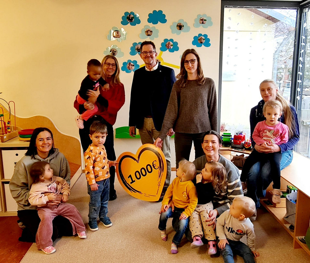 Das Bild zeigt einen Kindergartenraum, in dem mehrere Erwachsene und Kinder anwesend sind. Im Vordergrund ist ein herzförmige