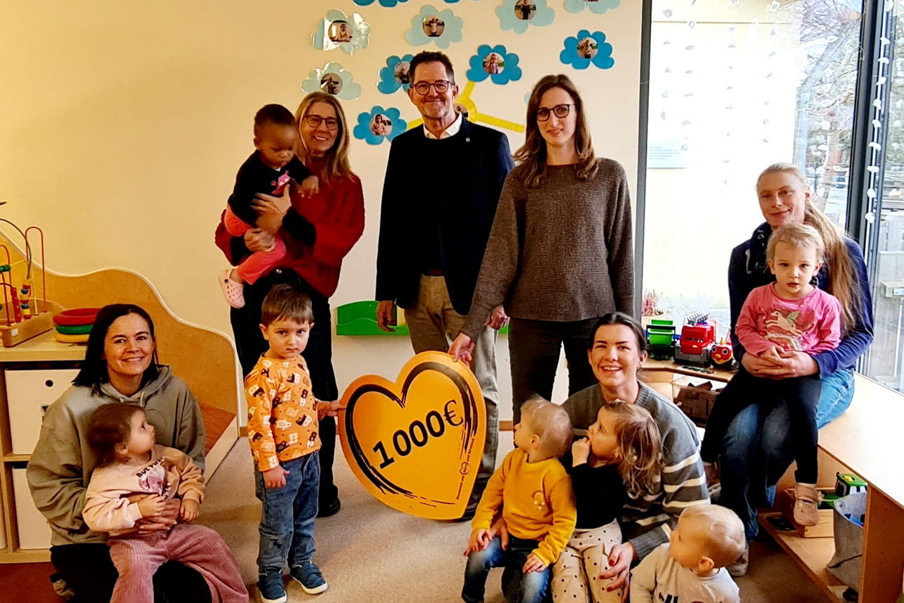 Das Bild zeigt einen Kindergartenraum, in dem mehrere Erwachsene und Kinder anwesend sind. Im Vordergrund ist ein herzförmige