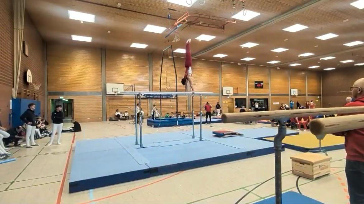 Turnhalle mit Turngeräten wie Barren und Matten, Personen stehen am Rand und beobachten.