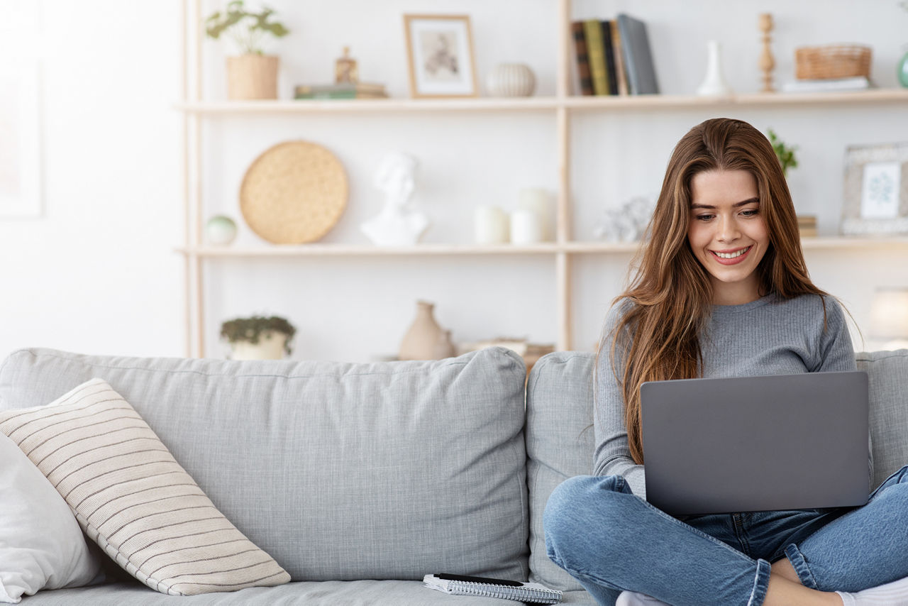 Lächelnde junge Frau, die von zu Hause aus arbeitet, auf der Couch sitzt und Laptop benutzt