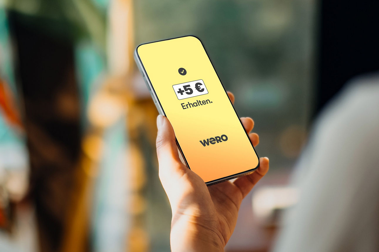 Eine Hand hält ein Smartphone und auf dem Display sieht man das Wero-Logo und den Schriftzug "+5 € Erhalten"