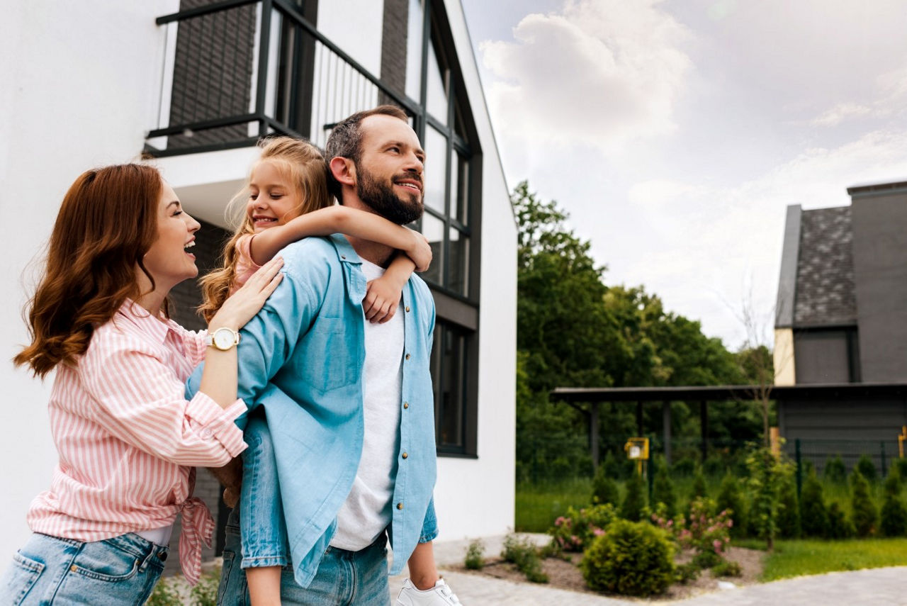 Eine fröhliche Familie steht vor einem modernen Haus. Das Kind sitzt auf den Schultern des Vaters, die Mutter steht daneben und lacht.