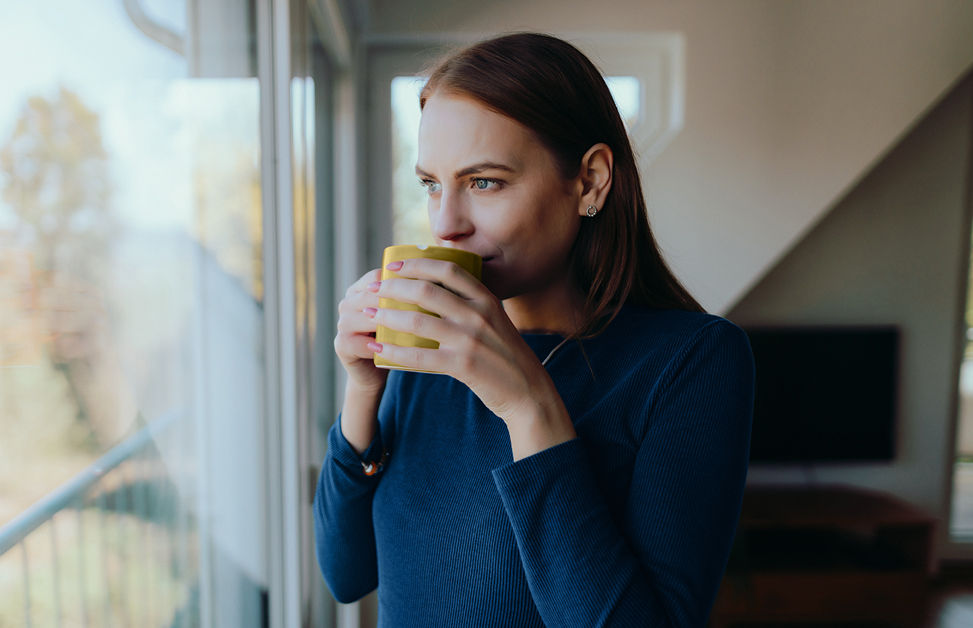 Frau steht mit Tasse in der Hand vorm Fenster