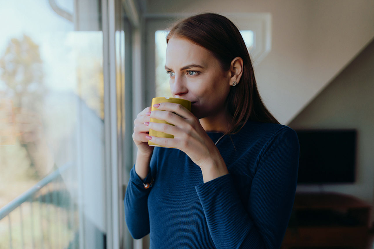 Frau steht mit Tasse in der Hand vorm Fenster