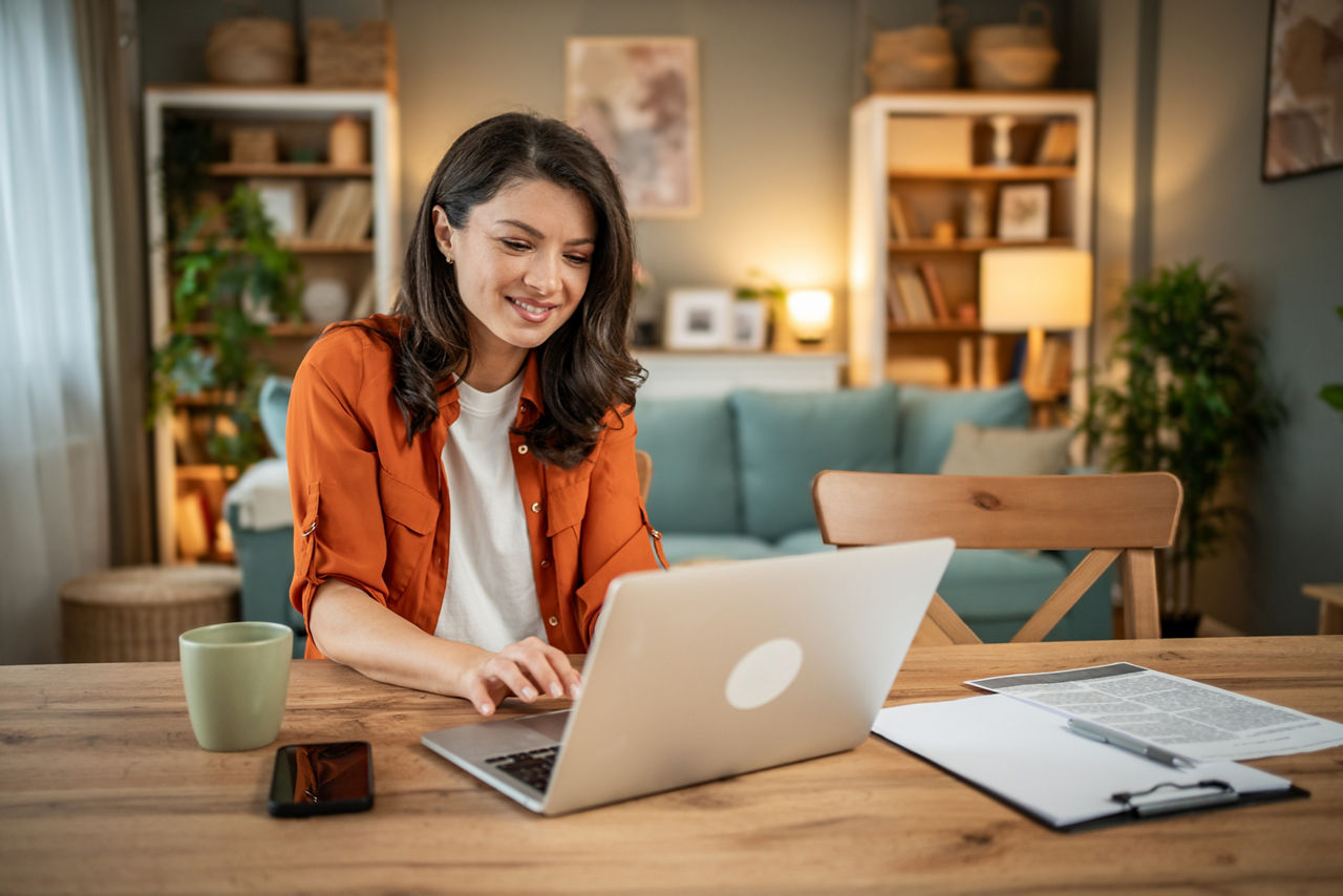 Frau mit oranger Jacke sitzt an Ticsh und arbeitet an Laptop