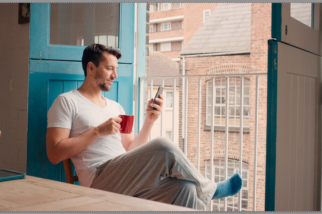 Junger Mann sitzt am Fenster mit Kaffee und seinem Smartphone in der Hand
