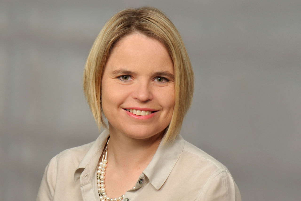 Nicole Mouzon hat blonde kurze Haare und  trägt eine helle Bluse mit einer Perlenkette