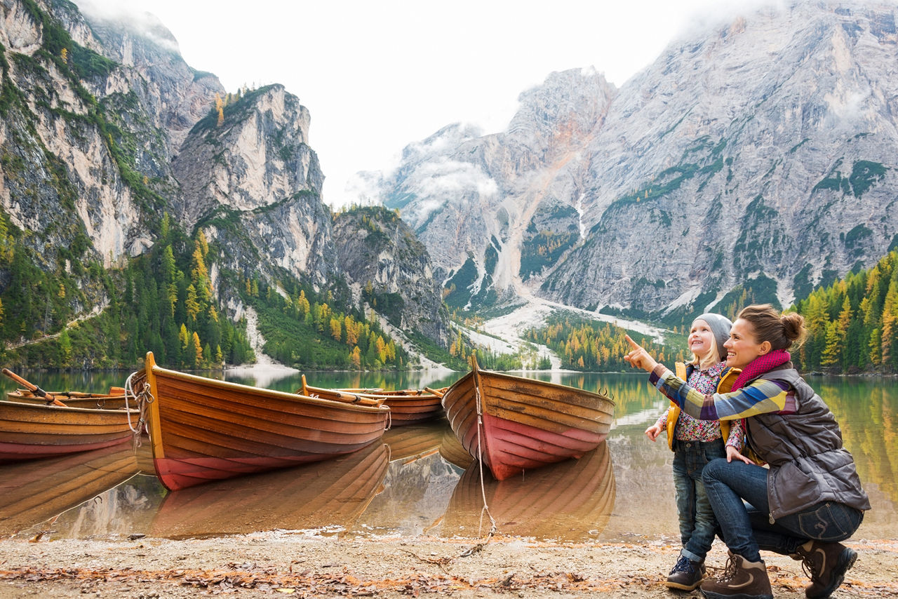 Eine lächelnde Mutter kniet neben ihrer Tochter und zeigt nach oben. Im Hintergrund schwimmen Holzboote auf dem Wasser, Herbstfarben, Dolomiten und Wald.