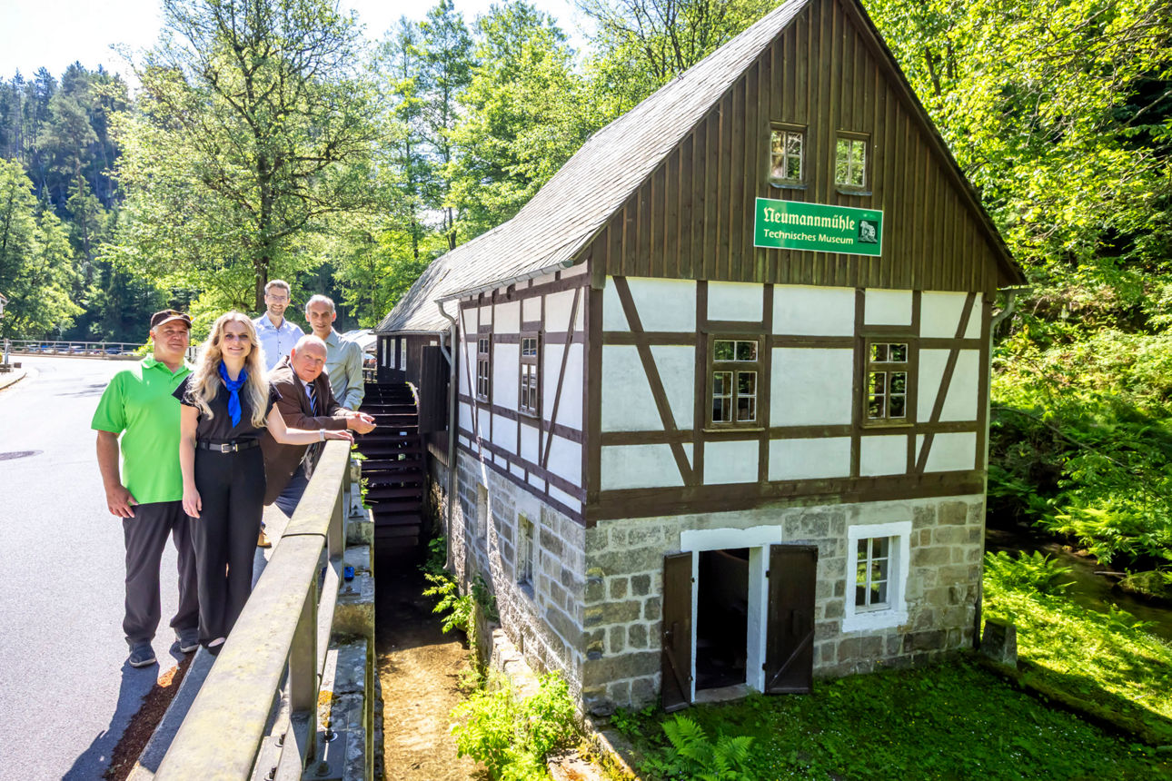 450 Jahre Neumannmühle – Als das Kirnitzschtal vom Klang der Sägen erfüllt war06.06.2026, Kirnitzschtal, Sebnitz, Neumannmühle Kirnitzschtal. Im Jahr 2026 erinnerte die traditionsreiche Anlage an ein außergewöhnliches Jubiläum: Vor 450 Jahren, am 2. September 1576, wurde die Neumannmühle erstmals urkundlich erwähnt. Was damals als einfache Brettmühle am Eingang des Großen Zschands begann, entwickelte sich zu einem bedeutenden Zeugnis der Wirtschafts- und Technikgeschichte der Sächsischen Schweiz. Dabei reichte die Geschichte vermutlich noch weiter zurück. Historiker vermuteten, dass bereits im 14. Jahrhundert an dieser Stelle eine kleine Schneidemühle gestanden hatte. Sicher belegt war jedoch erst die Erwähnung von 1576 in einem Verzeichnis des Amtes Hohnstein. Dort wurden an der Einmündung des Großen Zschands in das Kirnitzschtal zwei Brettmühlen genannt – eine davon war die heutige Neumannmühle.Mit dem Beginn der Holzflößerei auf der Kirnitzsch gegen Ende des 16. Jahrhunderts gewann die Mühle zunehmend an Bedeutung. Das Tal wurde zu einer wichtigen Verkehrsader für den Holztransport. Baumstämme aus den Wäldern der Hinteren Sächsischen Schweiz gelangten auf dem Wasserweg zu den Sägemühlen, wo sie zu Bau- und Nutzholz verarbeitet wurden. Die Neumannmühle profitierte von dieser Entwicklung und wurde über Generationen hinweg zu einem wichtigen Wirtschaftsbetrieb der Region. Besonders bemerkenswert war die Technik, die sich bis heute erhalten hat. Der älteste Teil der Mühle blieb über Jahrhunderte nahezu unverändert. Herzstück war ein Ein-Blatt-Sägegatter venezianischer Bauart – eine Konstruktion, die als technisches Meisterwerk ihrer Zeit galt. Während vielerorts historische Mühlentechnik verschwand, überdauerte sie in der Neumannmühle die Jahrhunderte nahezu im Originalzustand. Das mehr als vier Jahrhunderte alte Sägegatter gehört heute zu den bedeutendsten technischen Zeugnissen seiner Art in Sachsen.Einen weiteren Meilenstein erlebte die Mühle in den Jahren 1871 und 1872. Damals entstand ein Anbau für eine Holzschleiferei. Hier wurde Holzschliff hergestellt – jener Rohstoff, der die industrielle Papierproduktion revolutionierte. Täglich entstanden bis zu 300 Kilogramm Holzschliff, die in Papierfabriken der Umgebung weiterverarbeitet wurden. Die Neumannmühle schlug damit die Brücke zwischen traditionellem Handwerk und beginnender Industrialisierung. Angetrieben wurde die gesamte Anlage von der Kraft des Wassers. Das große Wasserrad setzte die Maschinen in Bewegung und lieferte eine Leistung von bis zu 18 Pferdestärken – lange bevor Elektrizität Einzug hielt. Für die Menschen jener Zeit war die Wasserkraft die Grundlage ihres wirtschaftlichen Erfolges.Nach dem Ende der Holzschliffproduktion im Jahr 1945 und der Einstellung des Sägebetriebes in den 1950er Jahren schien das Schicksal der Mühle zunächst besiegelt. Die Gebäude verfielen, die Technik rostete. Doch Heimatforscher, Denkmalpfleger und engagierte Bürger erkannten früh den unschätzbaren Wert der Anlage. Bereits in den 1960er Jahren begannen erste Bemühungen, die Mühle als technisches Denkmal zu sichern. Nach Jahren des Verfalls wurde sie ab den 1990er Jahren aufwendig restauriert. Seit der Gründung des Vereins „Schauanlage Neumannmühle e. V.“ im Jahr 1998 sorgten zahlreiche Ehrenamtliche dafür, dass die Geschichte der Mühle nicht in Vergessenheit geriet.So stand die Neumannmühle auch 450 Jahre nach ihrer ersten urkundlichen Erwähnung noch immer dort, wo Generationen von Müllern, Sägern und Handwerkern gearbeitet hatten. Als einzige Mühle im Kirnitzschtal hatte sie ihre ursprüngliche Gestalt weitgehend bewahrt. Sie war längst mehr als ein Museum. Sie war ein lebendiges Geschichtsbuch aus Holz, Stein und Eisen – ein Ort, an dem die Vergangenheit nicht nur erzählt, sondern erlebt werden konnte.Als sich am 6. Juni 2026 Förderer, Vereinsmitglieder, Freunde und Gäste im Technischen Denkmal versammelten, feierten sie deshalb nicht allein ein Jubiläum. Sie würdigten 450 Jahre Arbeit, Erfindergeist und Heimatgeschichte – und jene Menschen, die dafür gesorgt hatten, dass das alte Wasserrad im Kirnitzschtal auch für kommende Generationen nicht stillstehen würde.Foto: Marko Förster06.06.2026 © Marko FörsterWICHTIGE HINWEISE: Jeder Abdruck ist honorar- und umsatzsteuerpflichtig (7 % Ust.)! Die Weitergabe des Bildmaterials an Dritte ist verboten!