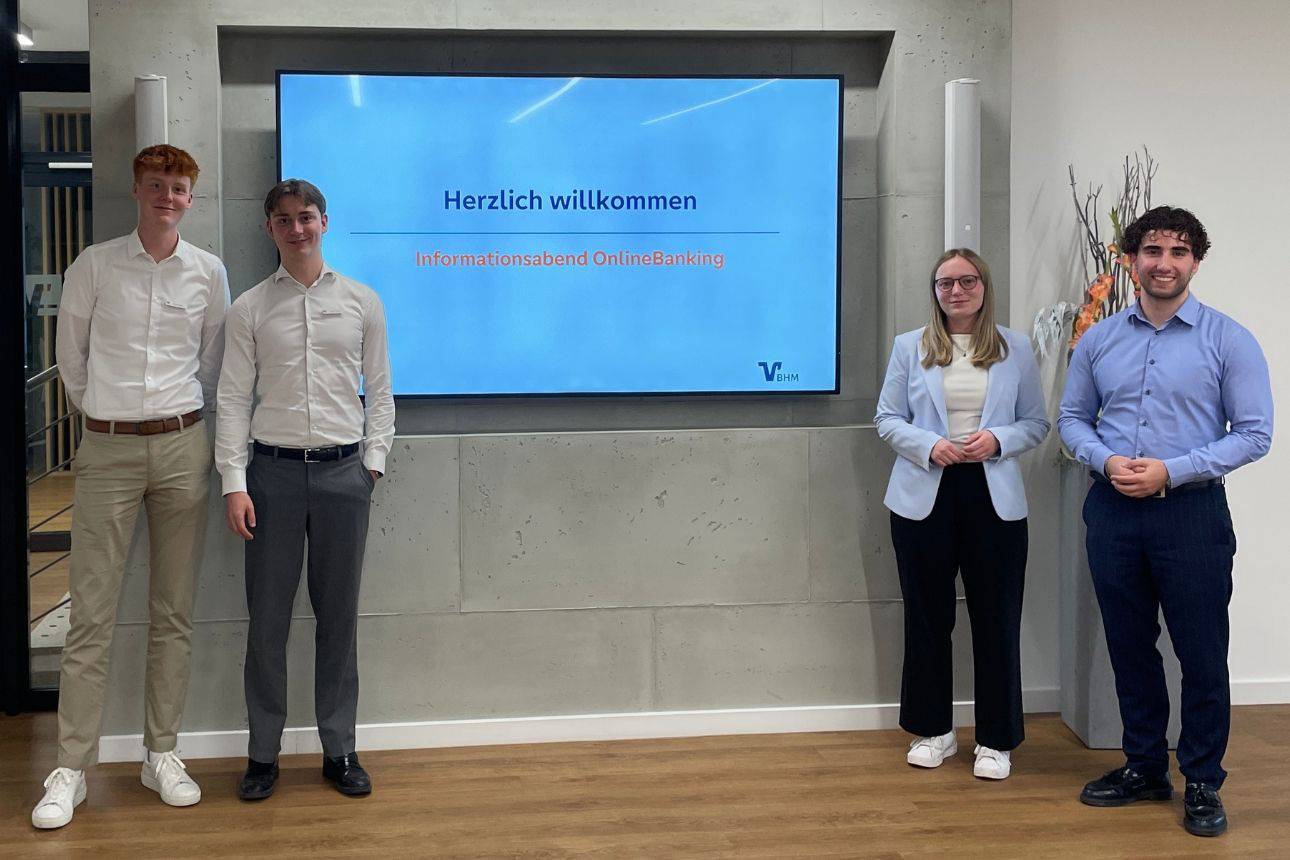 Vier Auszubildende stehen links und rechts neben einem Bildschirm mit der Aufschrift „Herzlich willkommen – Informationsabend OnlineBanking“. Der Bildschirm ist in der Hauptstelle der Volksbank in der Hohen Mark eG an einer Betonwand montiert. Unten rechts auf der Folie ist das Logo „VBHM“ (=Volksbank in der Hohen Mark eG) zu sehen.