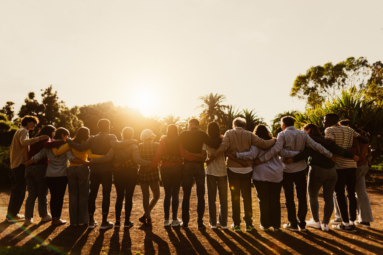 Abbildung zeigt eine Gruppe von Menschen jeden Alters, welche Arm in Arm in den Sonnenuntergang schauen