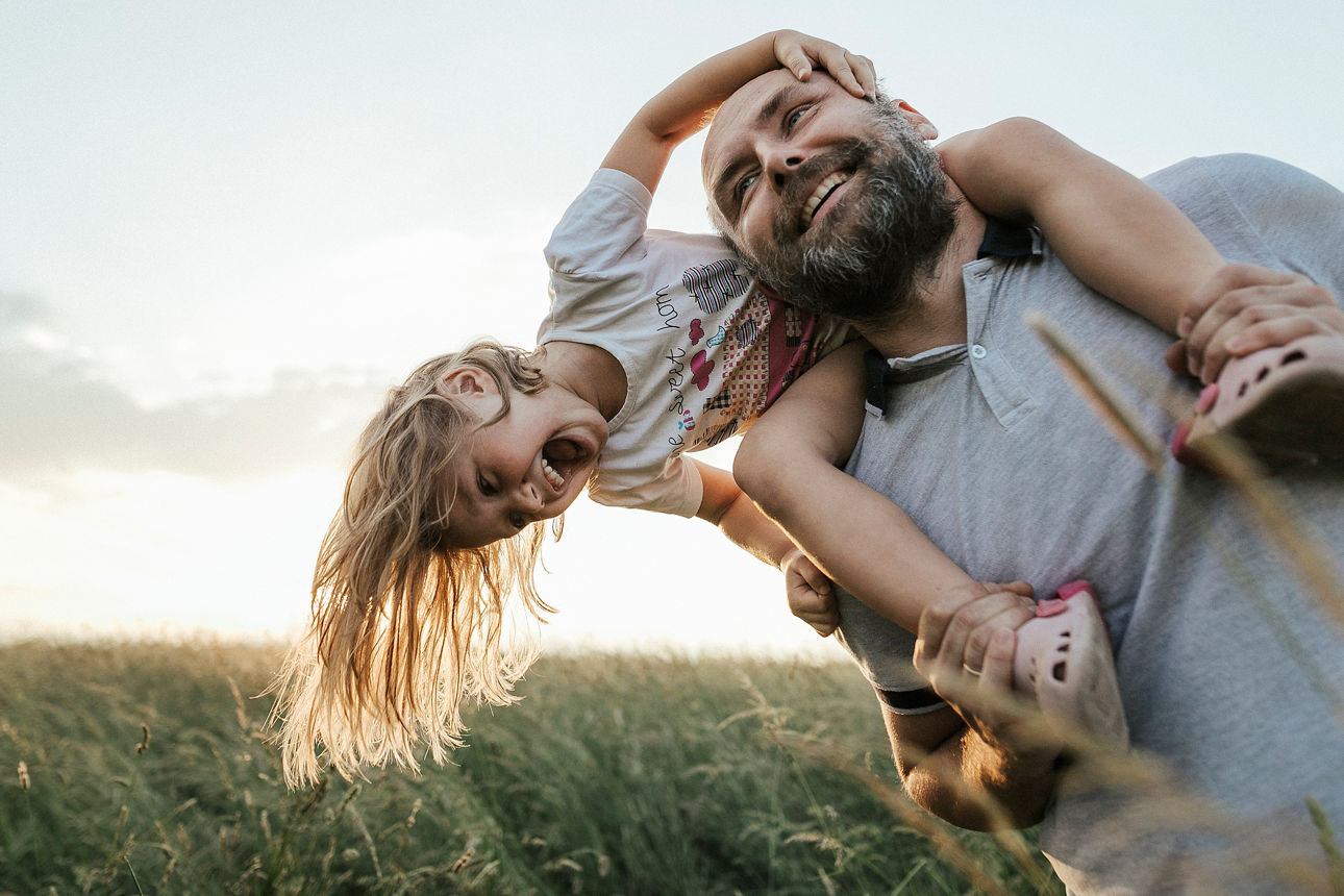 Ein Mann steht auf einer grünen Wiese und hat ein Mädchen auf den Schultern