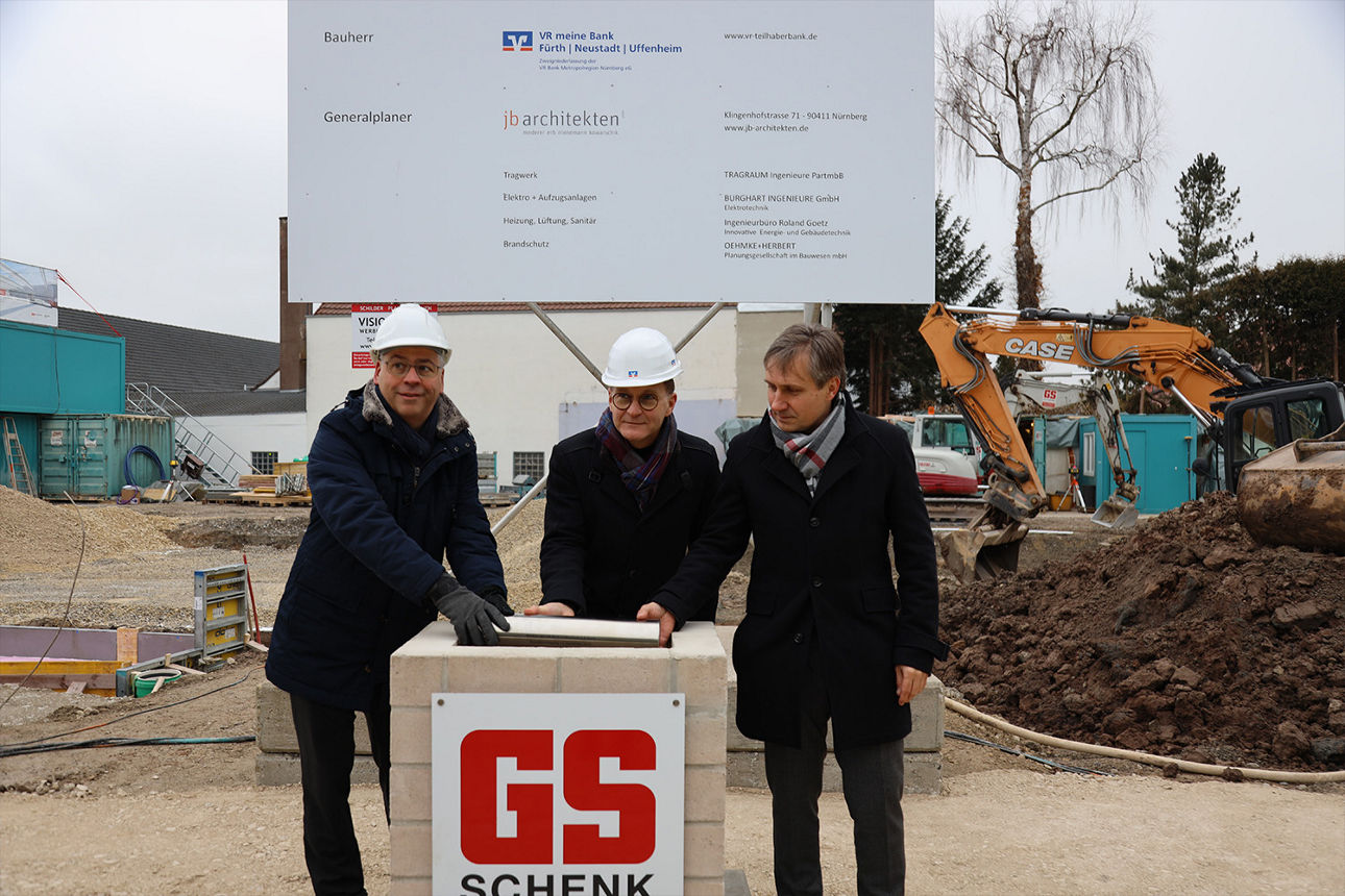 Drei Männer stehen vor einer Baustelle und halten den Grundstein