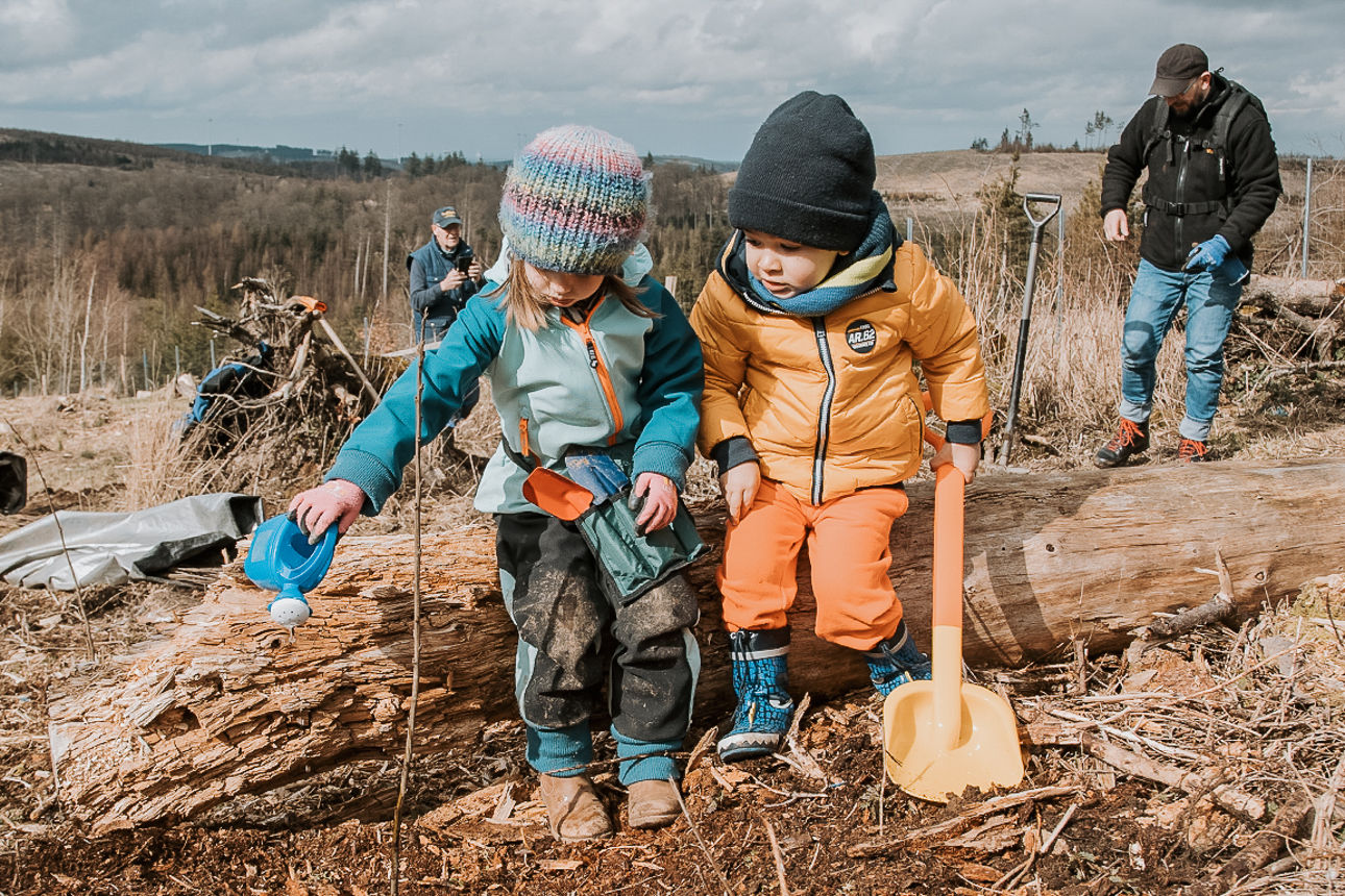 Zwei Kleinkinder mit Gießkanne und Schaufel bei einer Baumpflanzaktion.
