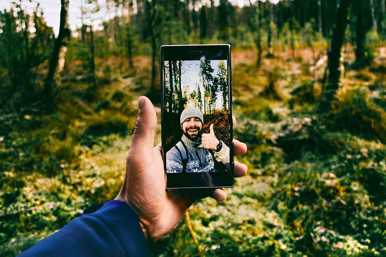 Mann hält im Wald ein Handy in der Hand, auf dem ein Selfie mit Daumen nach oben zu sehen ist