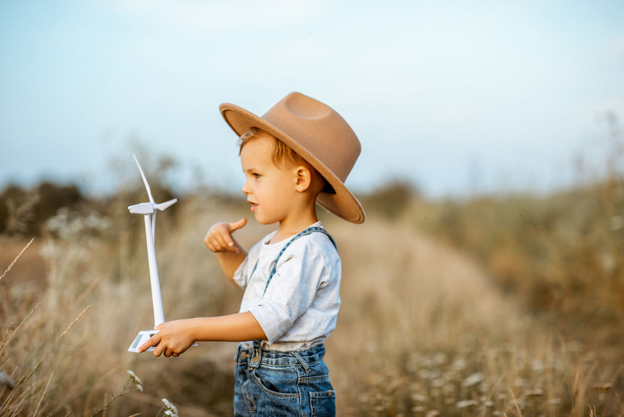 Kleiner Junge mit Cowboyhut steht auf einem Feld und hält Miniatur-Windrad in der Hand