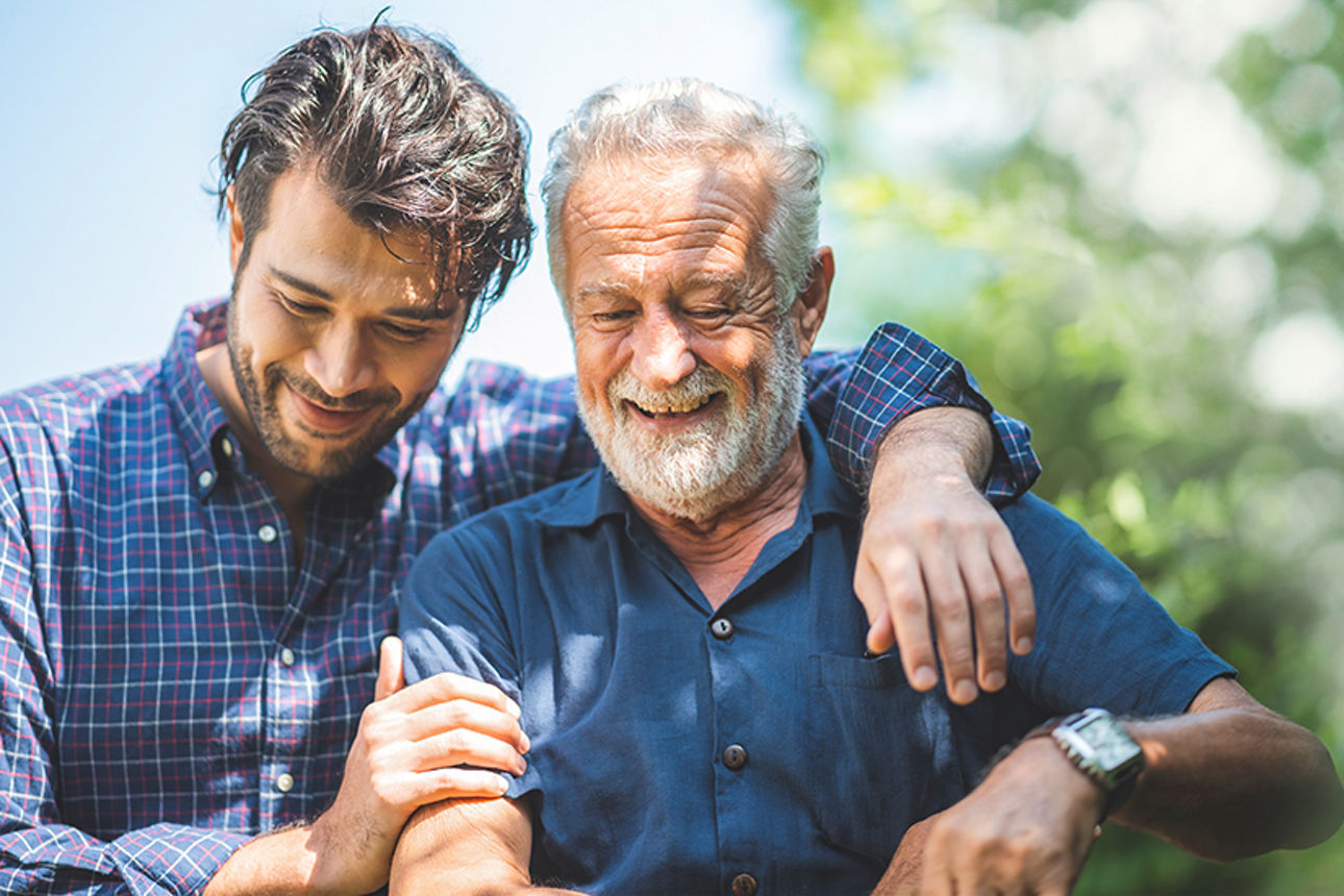 Ein älterer Mann und ein jüngerer Mann stehen im Park. Der jüngere Mann legt seinen Arm um die Schulter des älteren Mannes