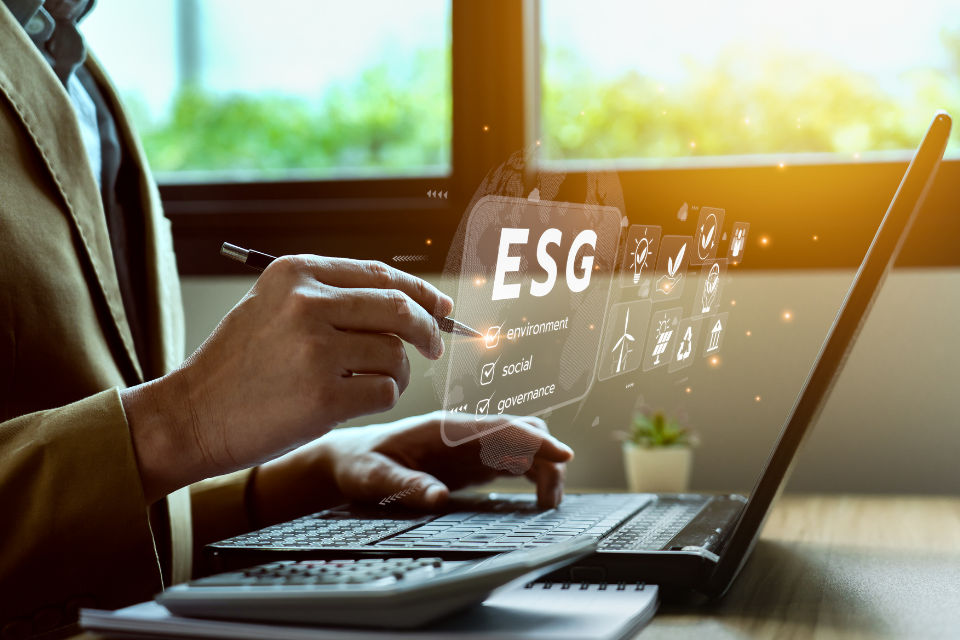 Eine Person am Laptop interagiert mit einem holografischen Display, das "ESG" für Umwelt, Soziales und Unternehmensführung zeigt.