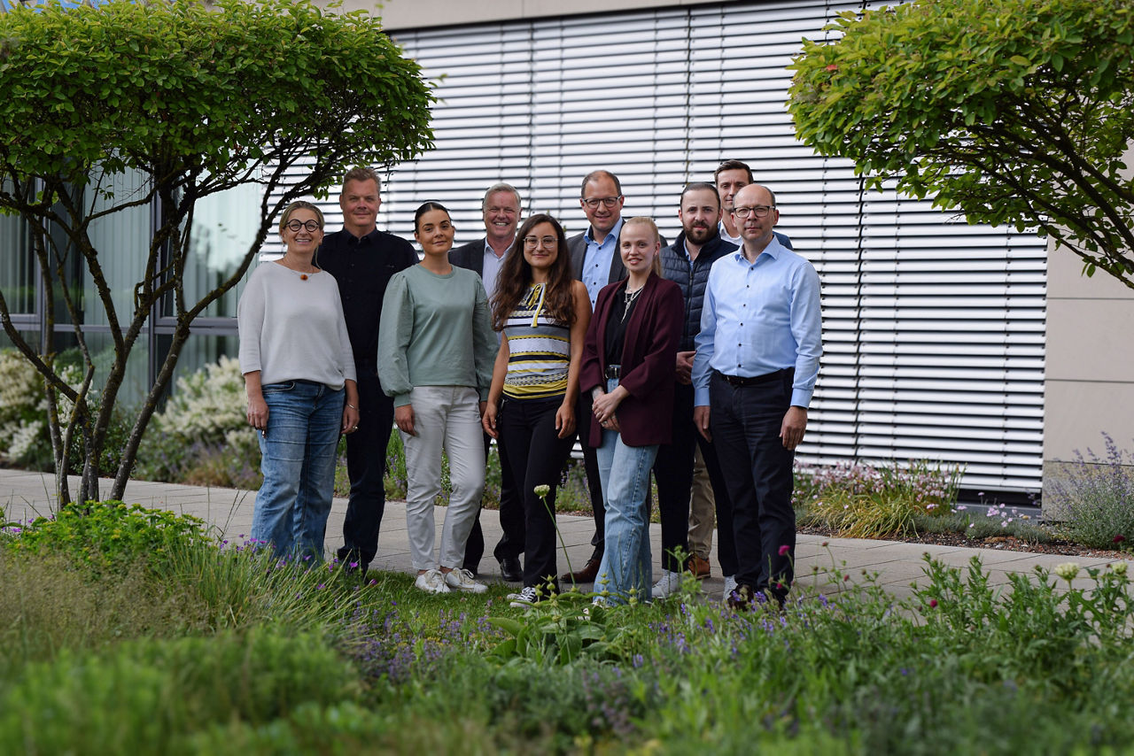 Gruppe von 10 Personen steht zwischen zwei Feigenbirnbäumen und blickt in die Kamera; davor  eine Wiese mit Wildblumen, dahinter die Jalousien eines großen Fensters
