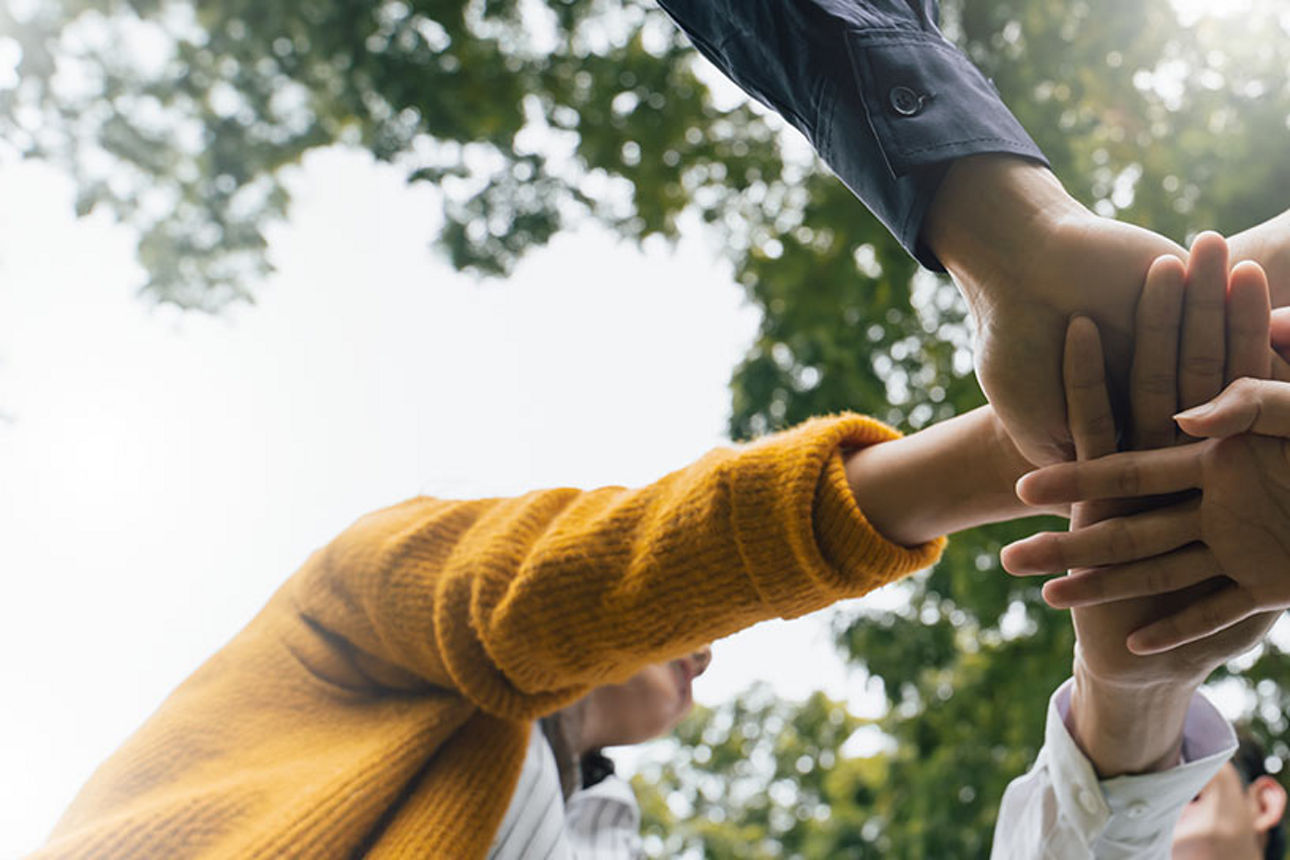 Mehrere Hände liegen übereinander in der Mitte; Menschen stehen im Freien unter Bäumen. Symbol für Zusammenarbeit, Gemeinschaft und Unterstützung.