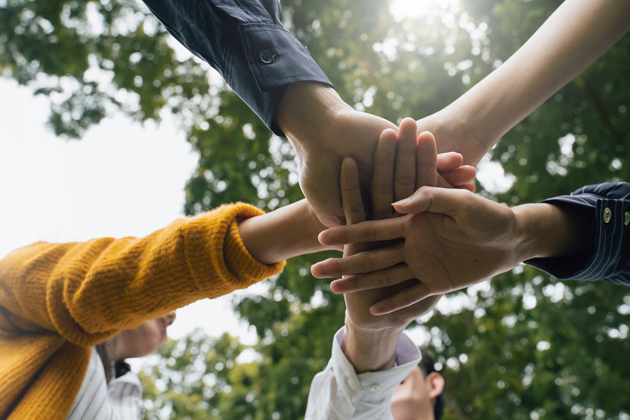Mehrere Hände liegen übereinander in der Mitte; Menschen stehen im Freien unter Bäumen. Symbol für Zusammenarbeit, Gemeinschaft und Unterstützung.