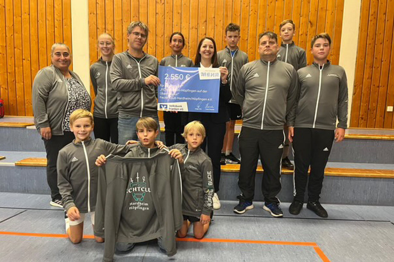 Eine Sportgruppe in Trainingsanzügen steht in einer Halle und hält einen großen Spendenscheck.