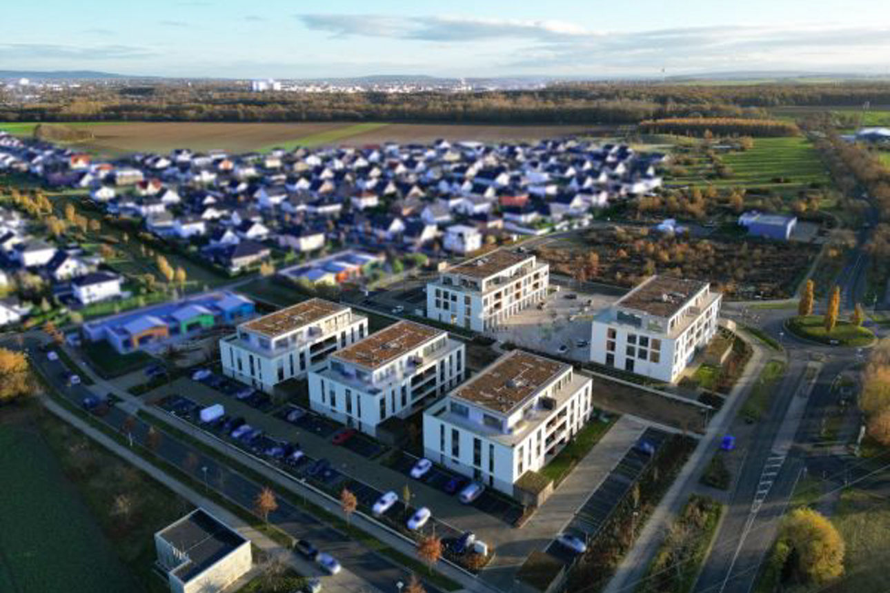 Luftaufnahme eines Neubaugebiets mit modernen Wohngebäuden, umgeben von Feldern, Straßen und einer angrenzenden Siedlung.